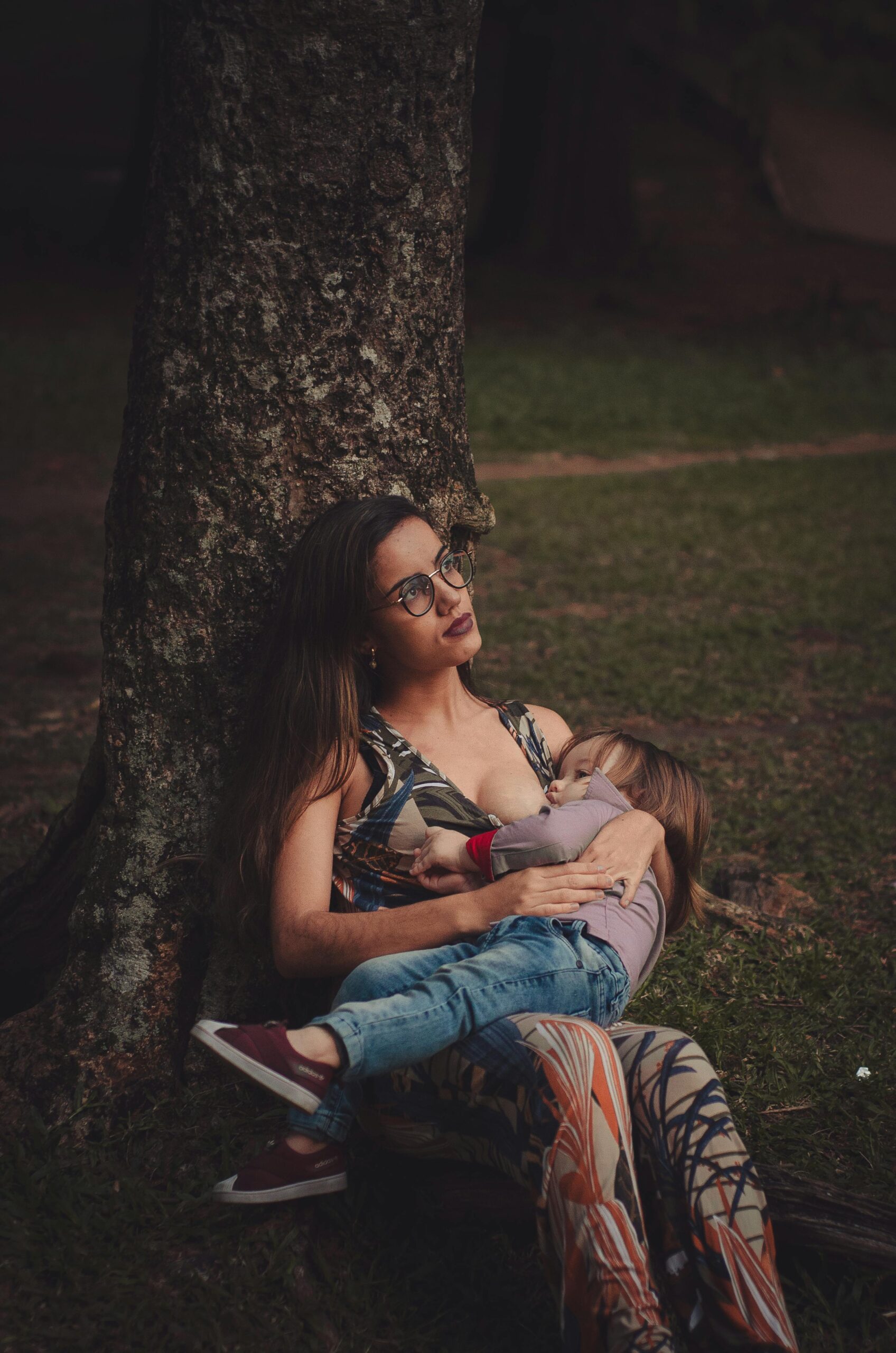 A mother lovingly embraces her child while sitting under a tree outdoors.